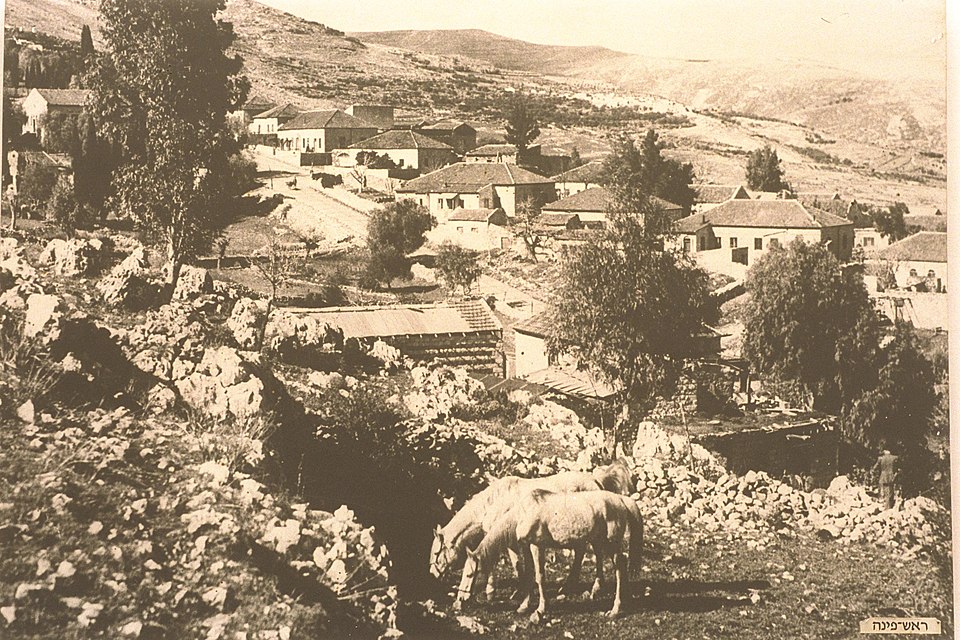 The moshava of Rosh Pina on a Galilee hillside in the late nineteenth century: tile-roofed settler houses among terraced rock, with horses grazing in the foreground.