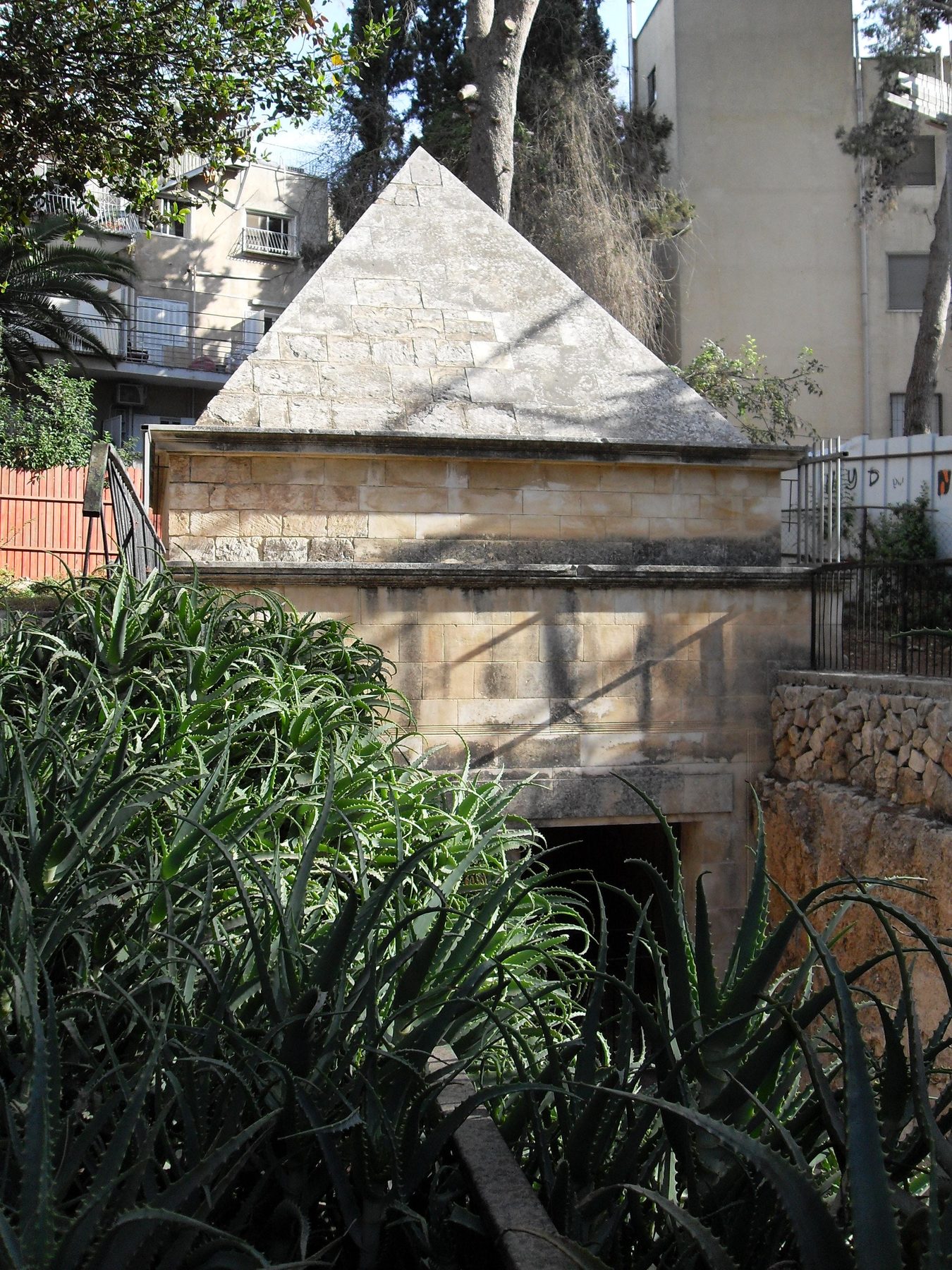 Hasmonean-era rock-cut tomb with pyramid-shaped stone roof and multi-layered porch facade, surrounded by modern Jerusalem buildings and vegetation.