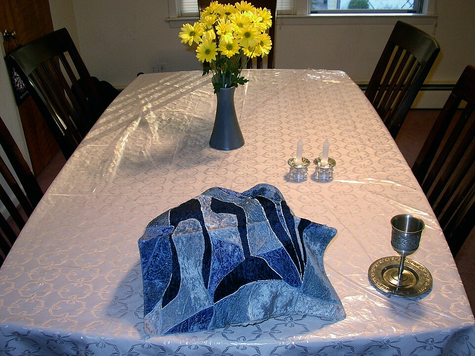 A Shabbat table set before sundown: covered challah, two candlesticks, a kiddush wine cup and flowers.