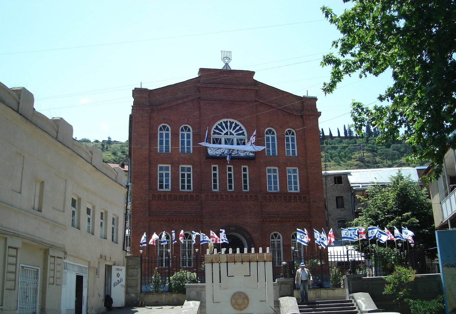 The red-brick Great Synagogue of Tbilisi, with a gabled facade topped by a menorah finial, Georgian and Israeli flags, and a Star-of-David monument in the forecourt