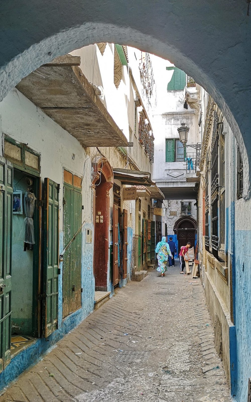 Narrow alley in Tetouan's mellah, with white-and-blue walls, old shopfront doors, and figures walking through the old Jewish quarter