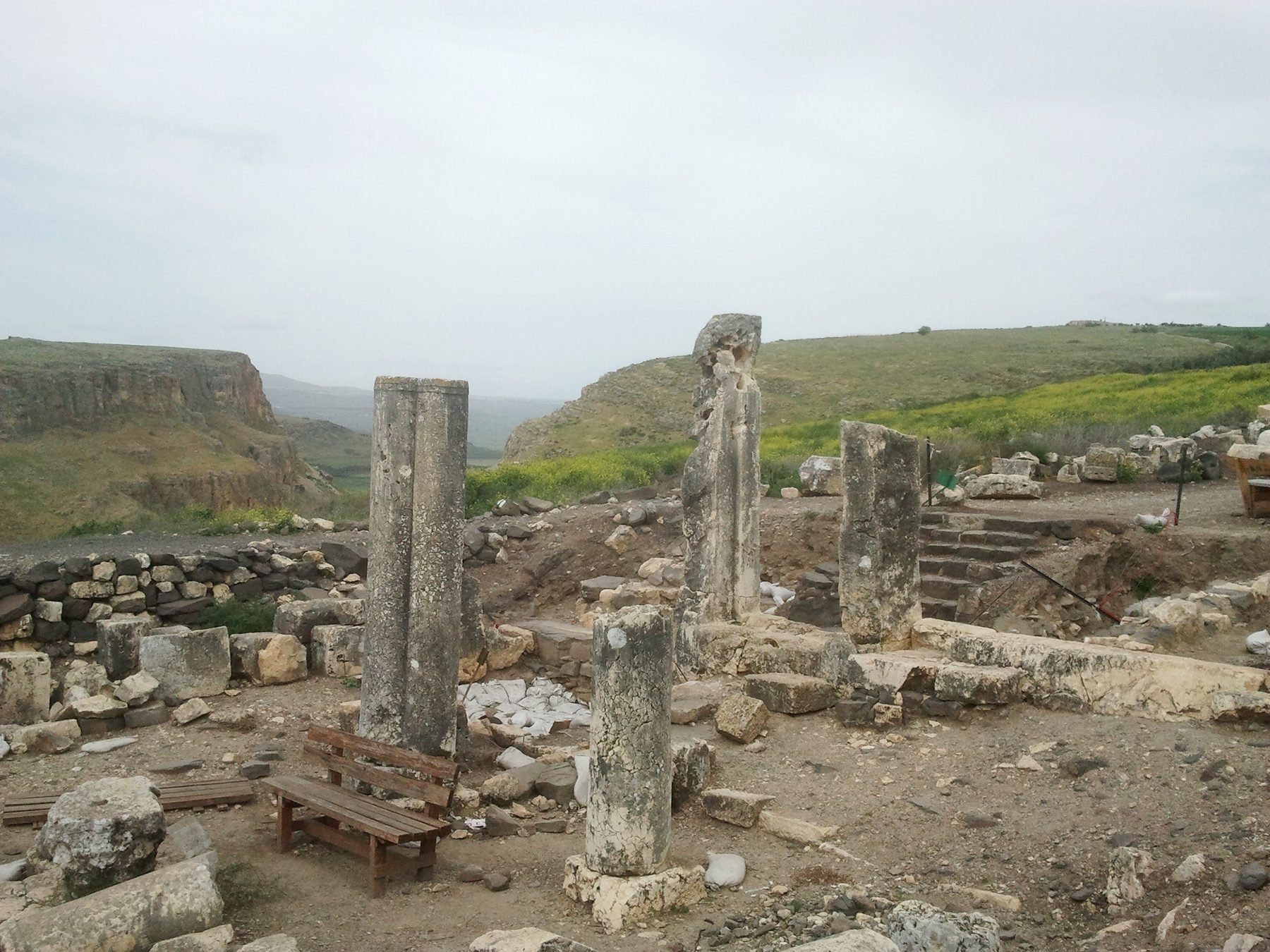Standing stone columns and foundation courses of the ancient Arbel synagogue beneath the basalt cliffs, with the Sea of Galilee visible in the distance.