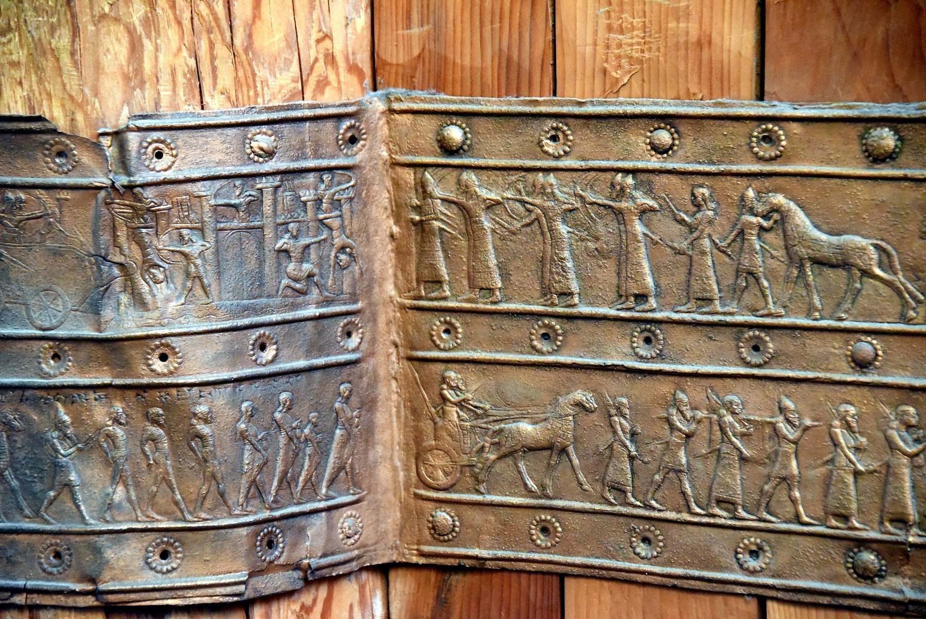 A bronze band from the Balawat Gates showing rows of Assyrian soldiers, chariots and captives in repoussé relief, with decorative rosette patterns framing the scenes.