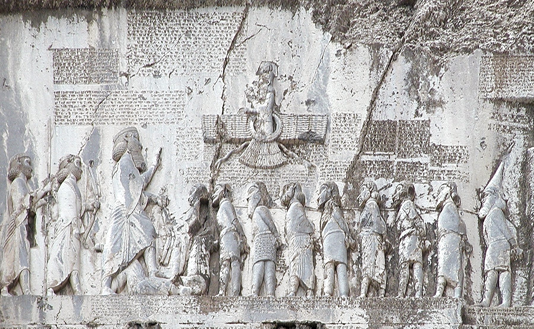 The Behistun Inscription relief carved high into a limestone cliff face, showing Darius I with foot planted on a defeated figure and facing a line of bound captives, with cuneiform text and a winged symbol above.