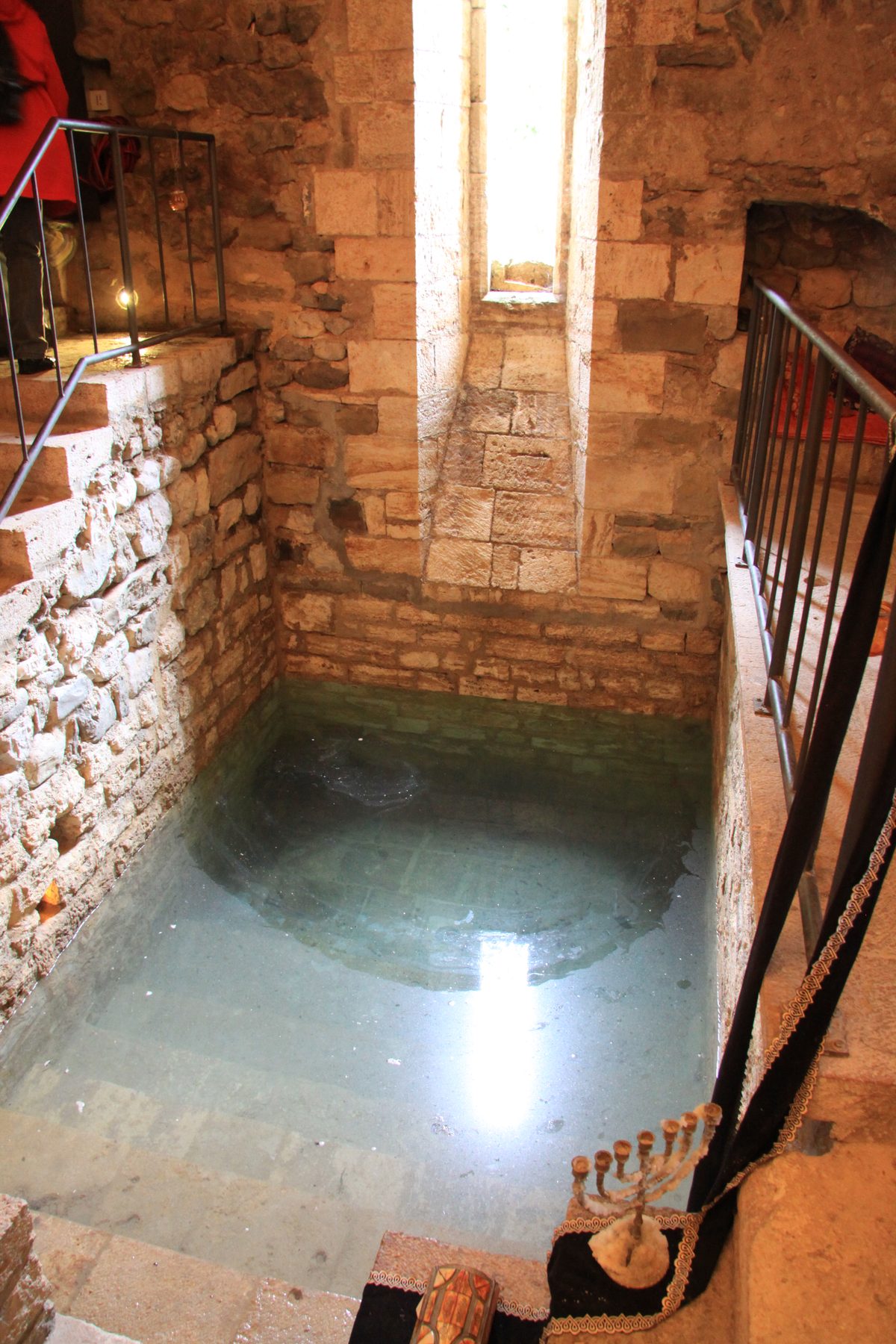 The stone-lined interior of the Besalú mikveh, showing the vaulted chamber walls and stepped descent to the water at the base, with metal railings on either side.