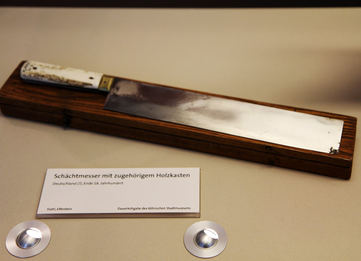 A long slender steel knife with a wooden handle laid on a dark surface.
