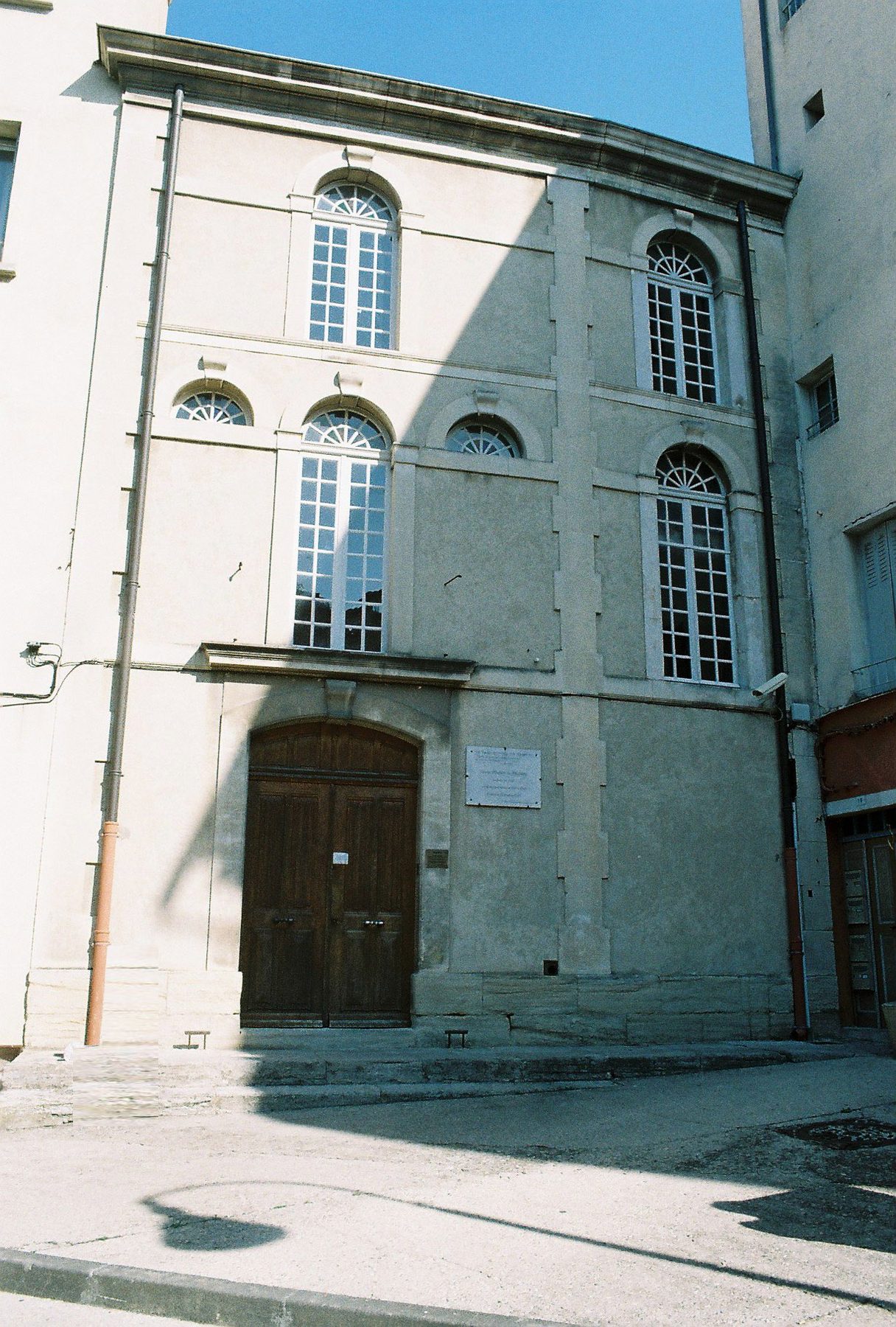 Stone facade of the Carpentras Synagogue with multiple rows of arched windows and a dark wooden entrance door.