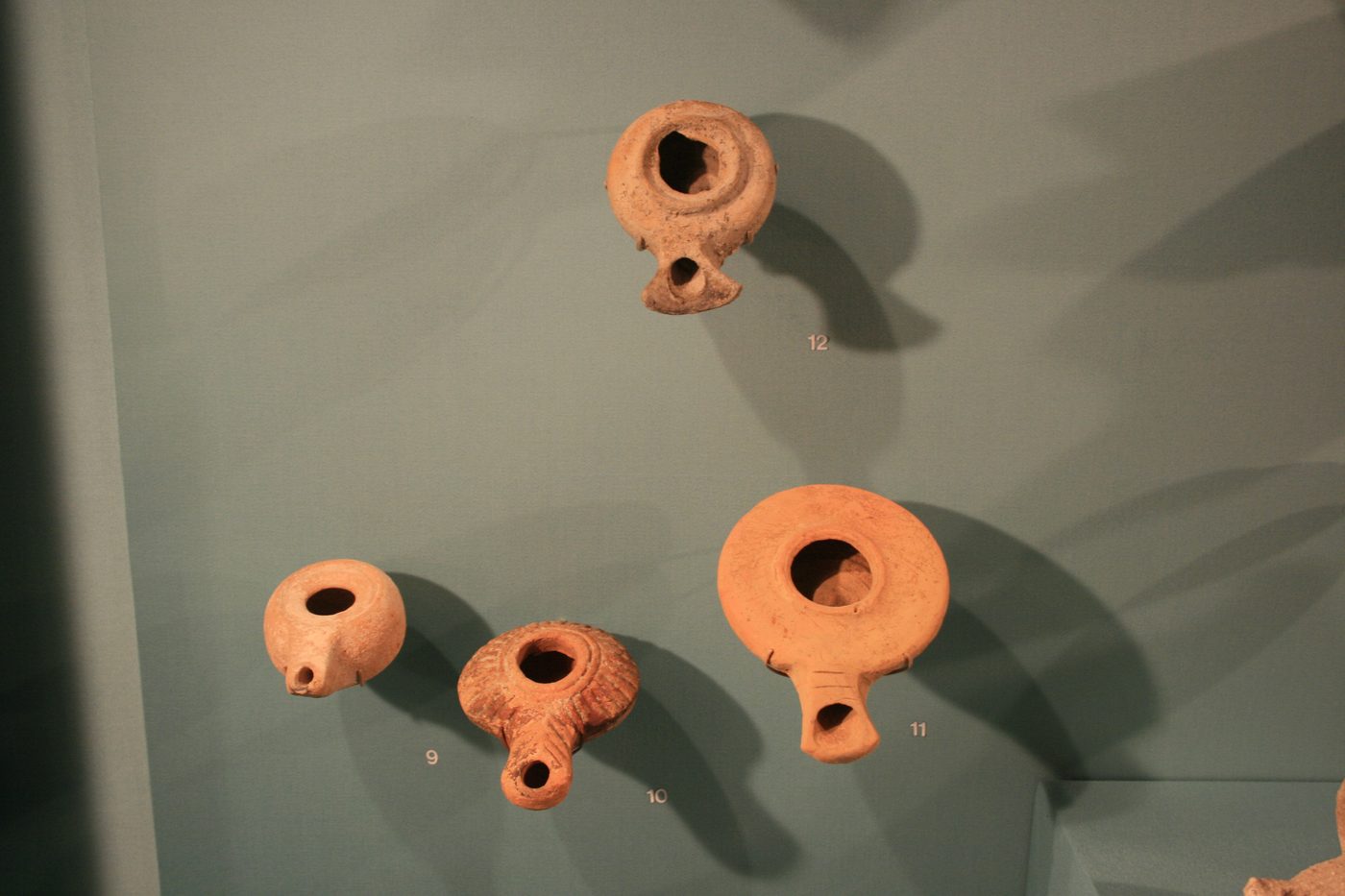 Four clay oil lamps mounted on a museum wall, numbered 9-12: two Hellenistic wheel-made lamps and two Herodian disc lamps with pinched nozzles, from the Levant