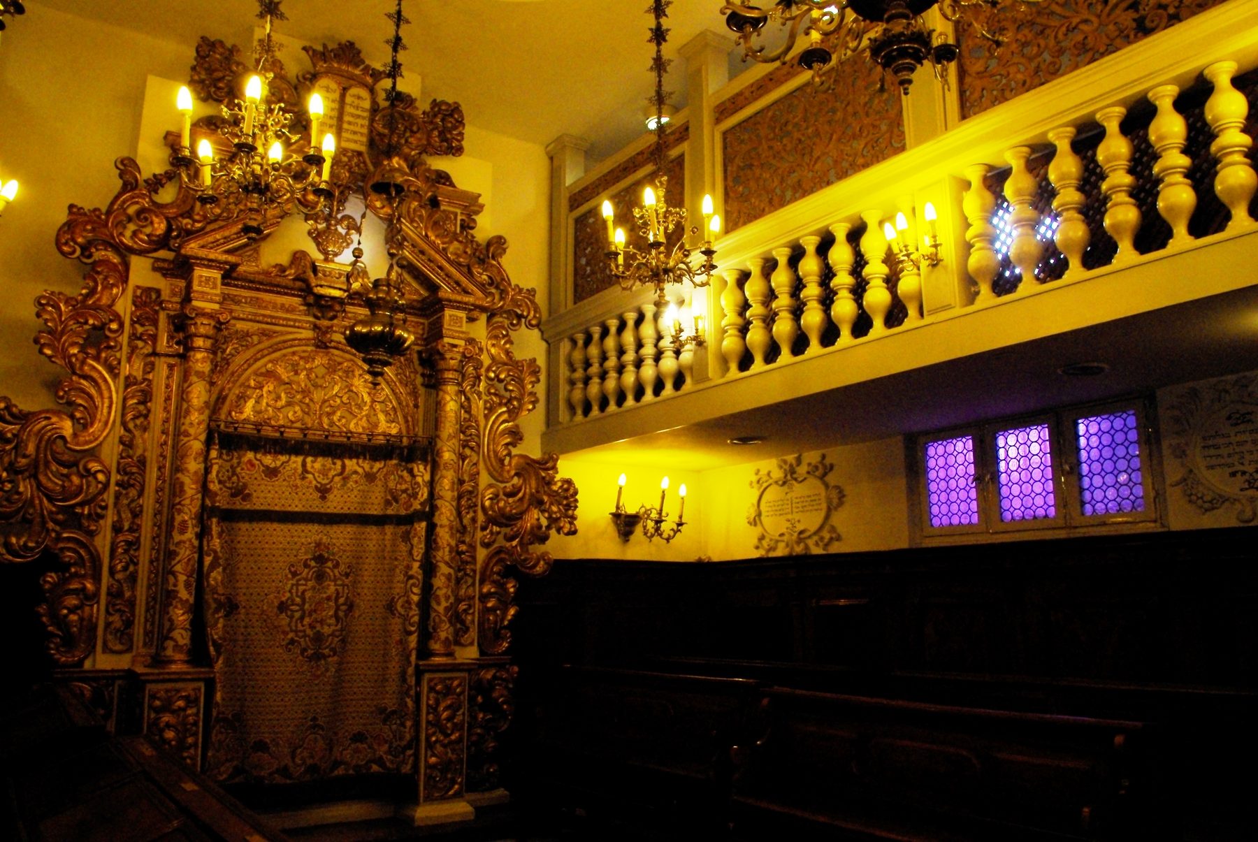 The ornate interior of the Conegliano synagogue, showing its gilded carved ark, raised bimah, wooden benches, and decorative plasterwork in the Rococo style.