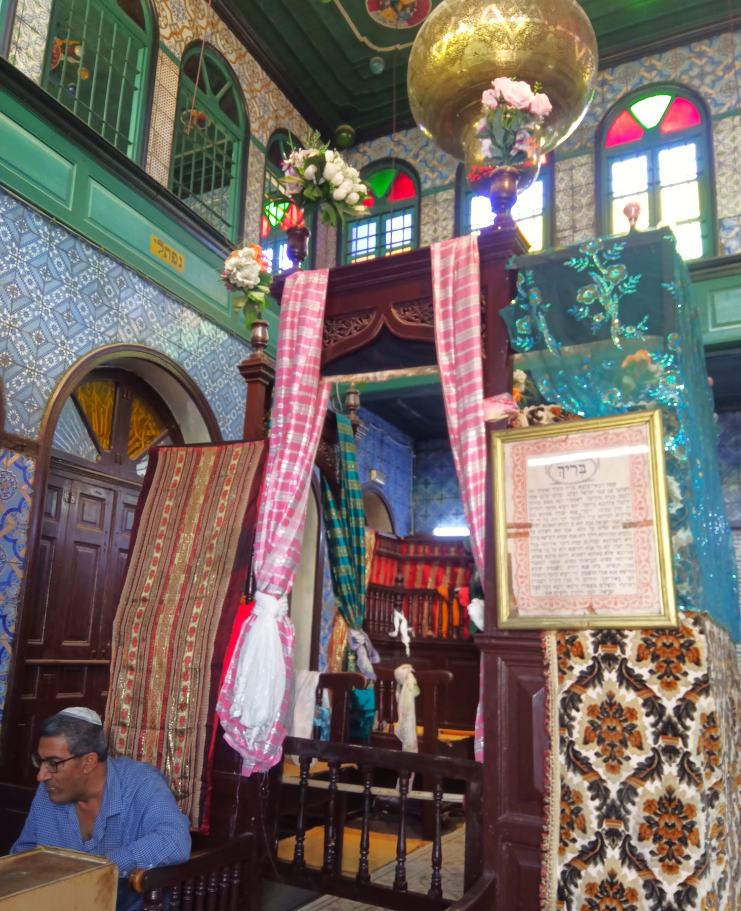 The ornate interior of the El Ghriba Synagogue, featuring blue tilework, stained-glass windows, and an elevated bimah with pink draped fabric