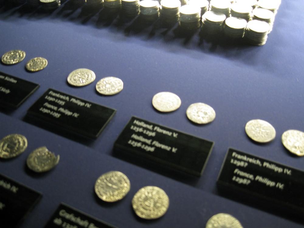Rows of medieval silver coins from the Erfurt Treasure laid out on a dark museum mount with printed labels naming their mints.
