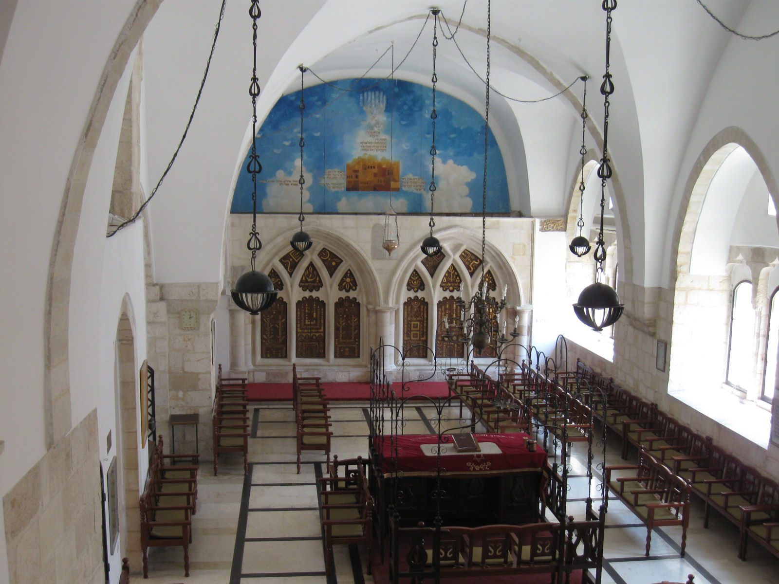 Interior prayer hall with stone barrel vaults, ornate ark, wooden benches, and turquoise decorative tilework with hanging oil lamps.