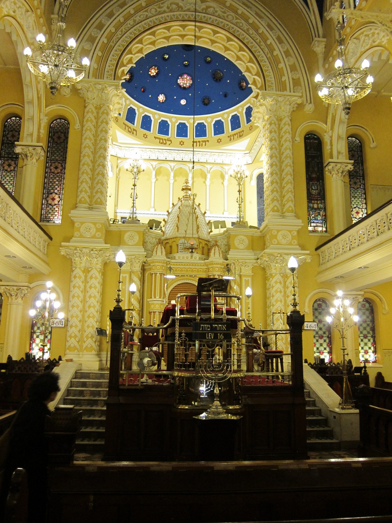Interior of The Great Synagogue's bimah, showing the raised prayer platform with the ark, ornate cream and gold wall decoration, crystal chandeliers, and the distinctive blue dome ceiling adorned with gold stars.