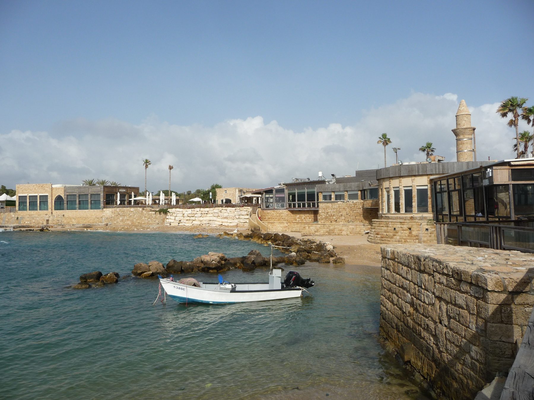 Stone remains of Herod's harbour at Caesarea Maritima, showing ancient breakwater structures in the Mediterranean with modern buildings and a mosque minaret on the coast.