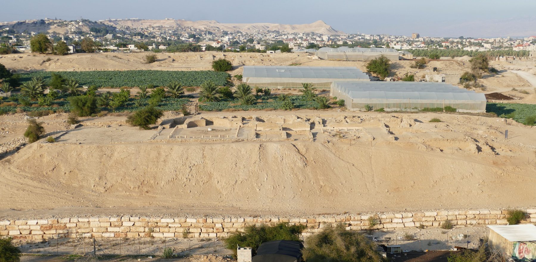 Excavated stone foundations and walls of the Hasmonean palace at Tulul Abu el-Alayiq near Jericho, with modern city and greenhouses visible beyond.