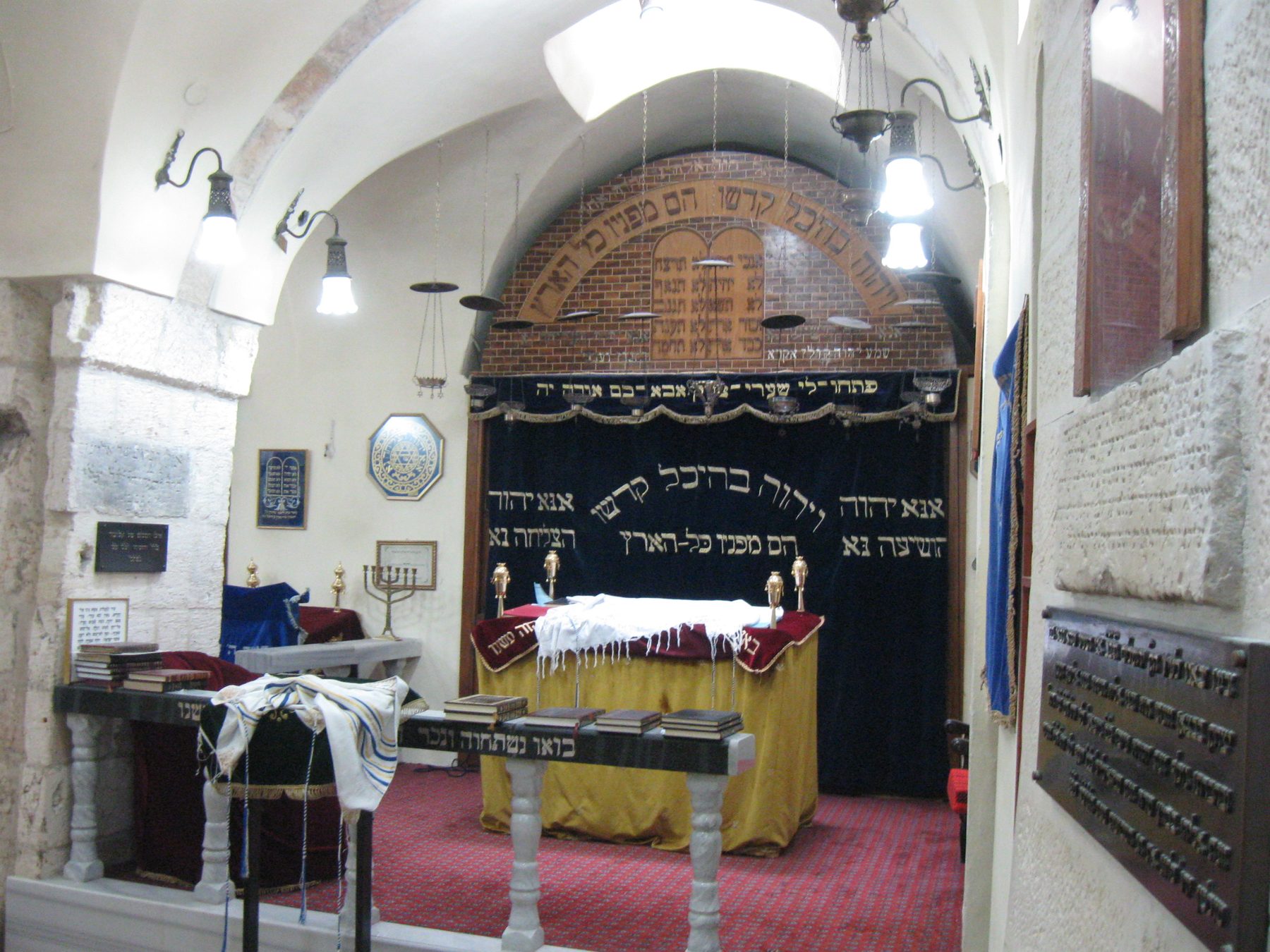 Interior of the sunken Karaite kenesa prayer hall showing the brick apse with Hebrew inscriptions, barrel-vaulted ceiling with skylights, burning candles, and religious furnishings.