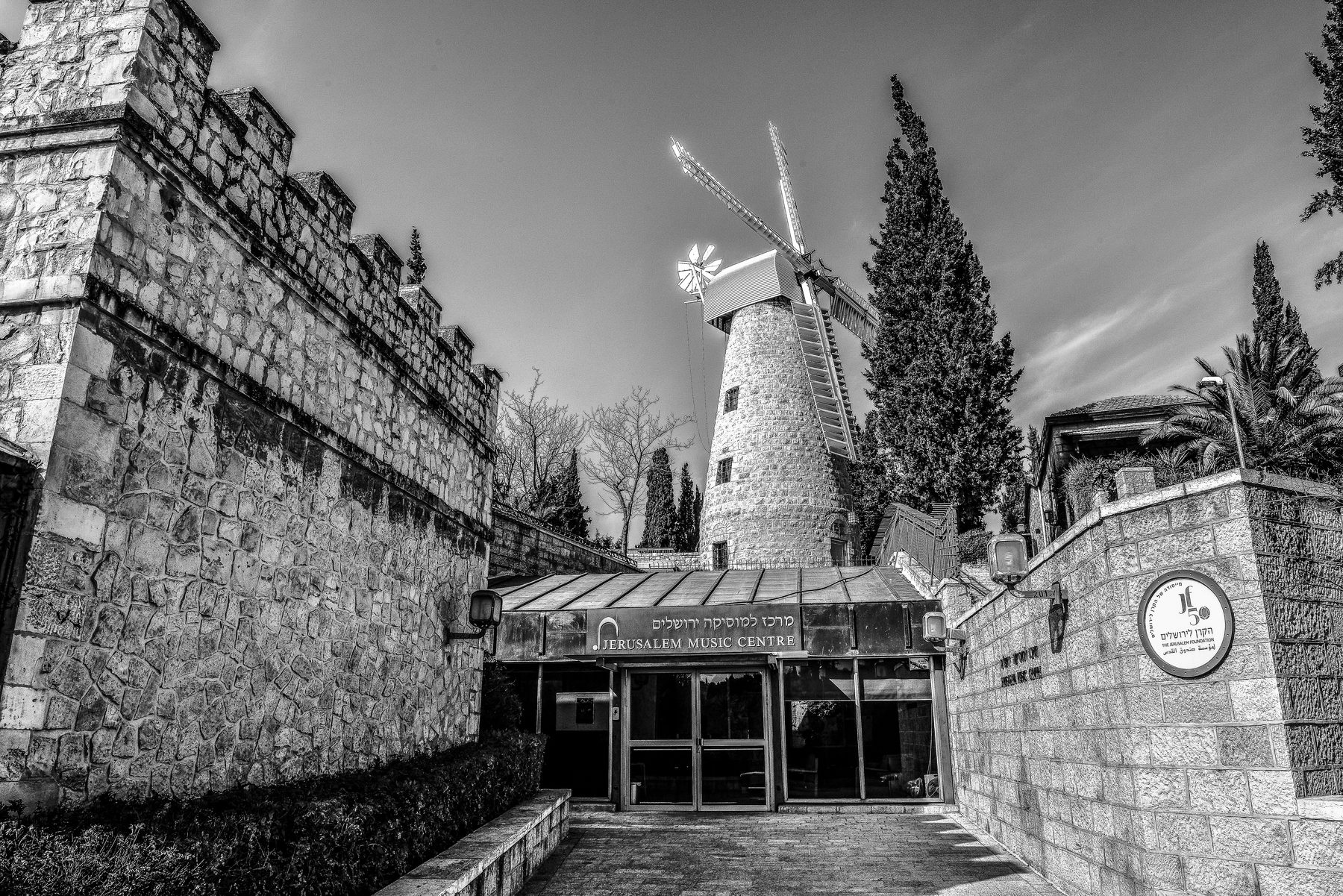 Stone windmill with rotating sails at Mishkenot Sha'ananim, Jerusalem, with cypress trees and stone buildings.