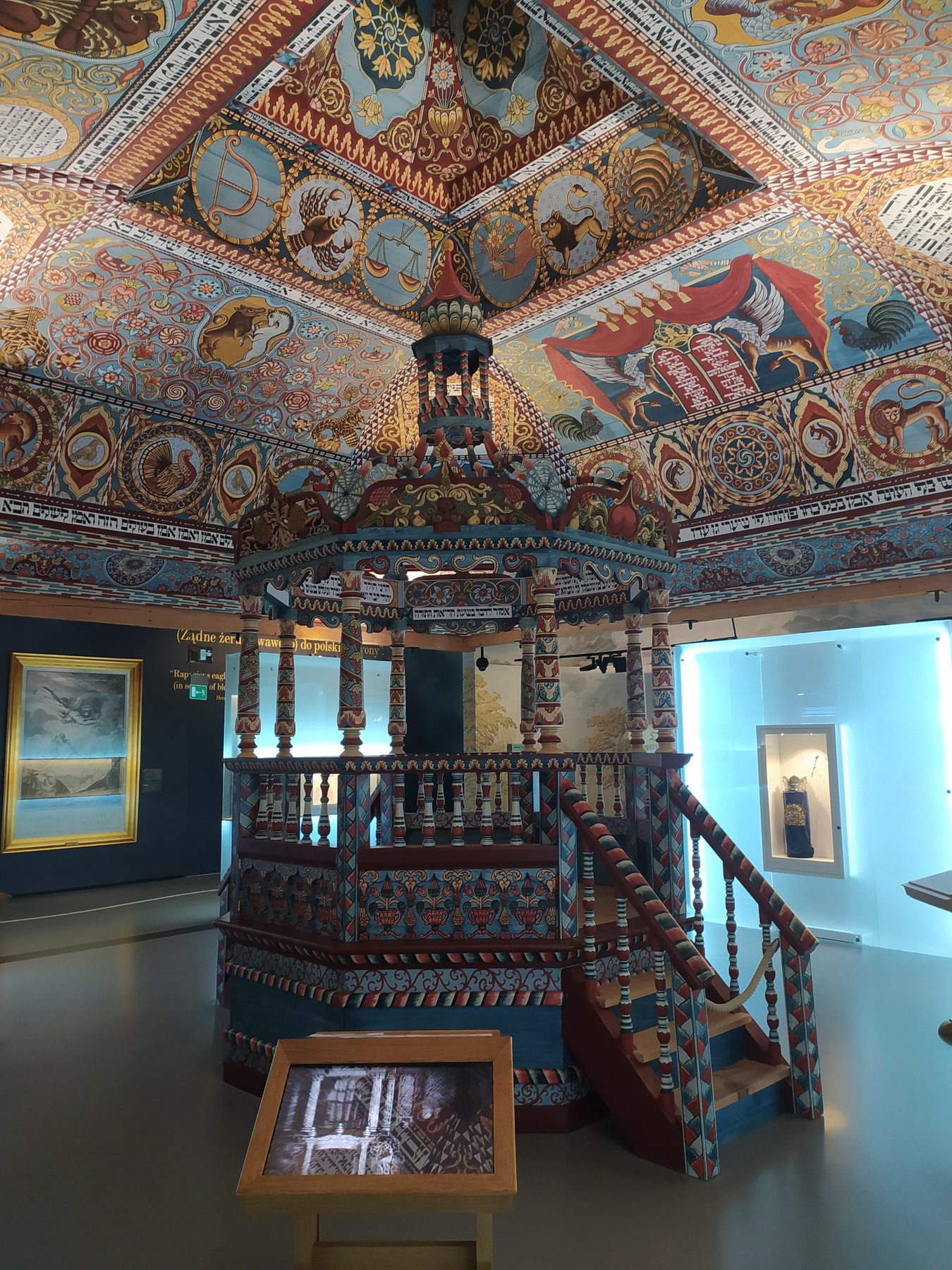 Reconstructed interior of the Gwoździec synagogue, showing the painted wooden ceiling with zodiac signs, lettering and ornamental patterns, and the tiered wooden bimah, at POLIN Museum.
