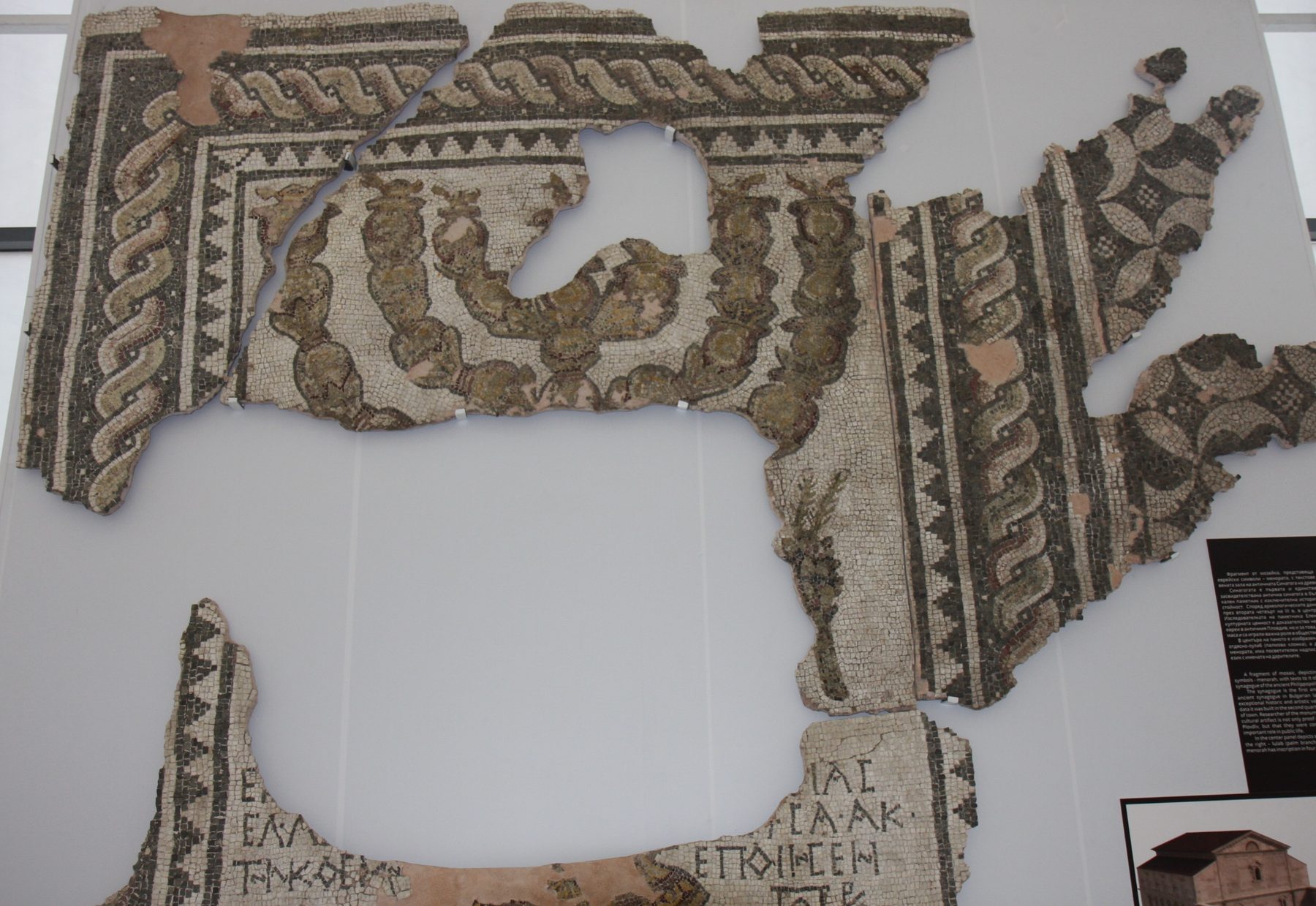 Fragmentary ancient mosaic floor featuring a menorah and geometric patterns, displayed on a museum mount.