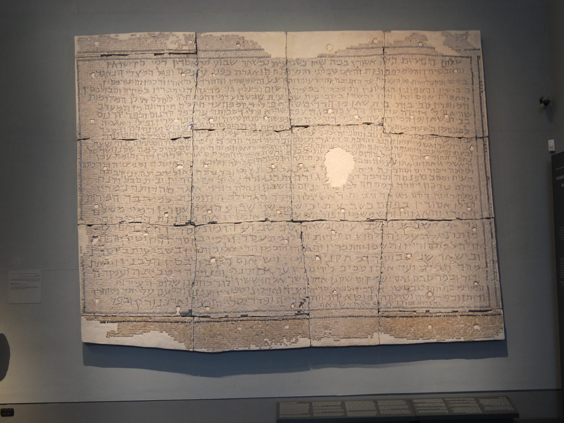 Ancient Hebrew mosaic floor inscription with multiple fragmented tile sections bearing Hebrew text, displayed in a museum setting.