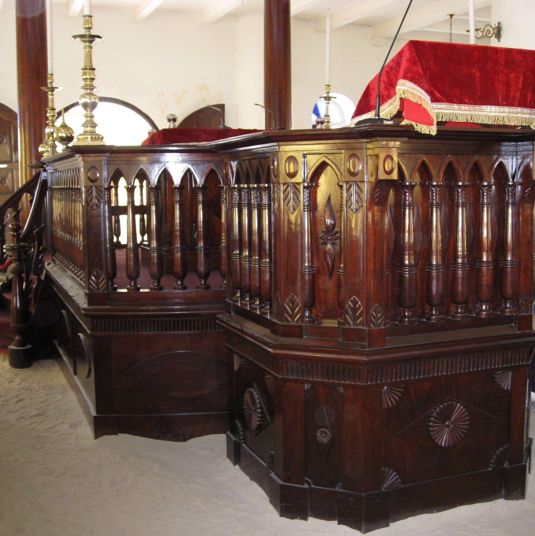 The interior sanctuary of the Snoa at Mikve Israel-Emanuel, showing the wooden bimah with ornate carved railings, sand floor, and brass fixtures.