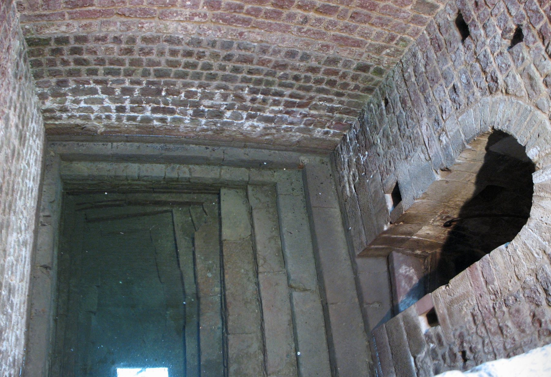 A stone stairwell with Romanesque arched brick ceiling descending through bedrock to a vaulted groundwater chamber, the interior of the Speyer mikveh.