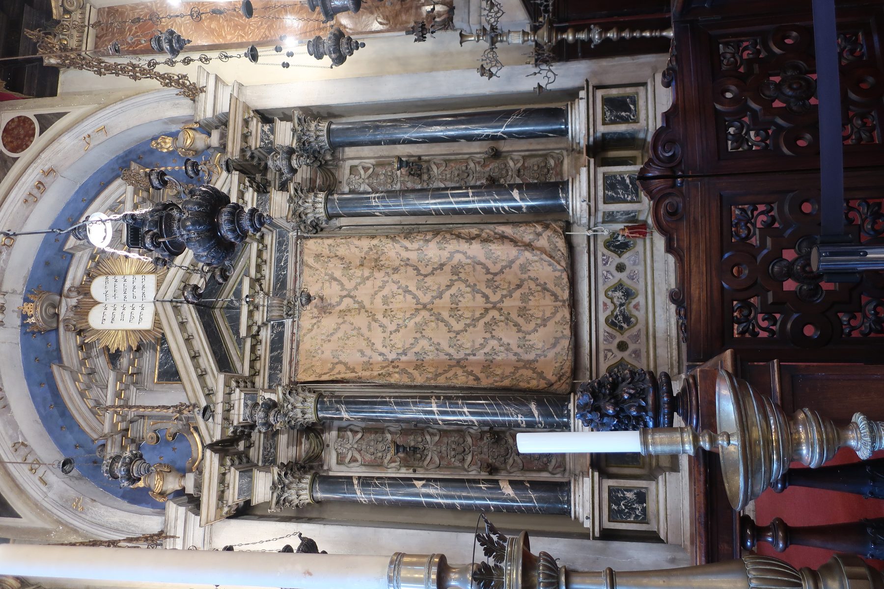Ornate interior of one of the Venice Ghetto synagogues viewed from above, showing gilded ceiling, multiple chandeliers, an ornamental Torah ark with Hebrew text panel, and decorative wooden panels.