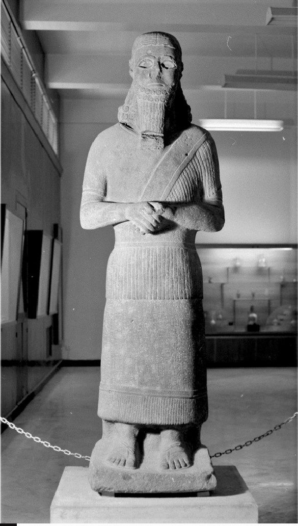 Basalt statue of a bearded ruler in a fringed robe with folded hands, on display in a museum gallery