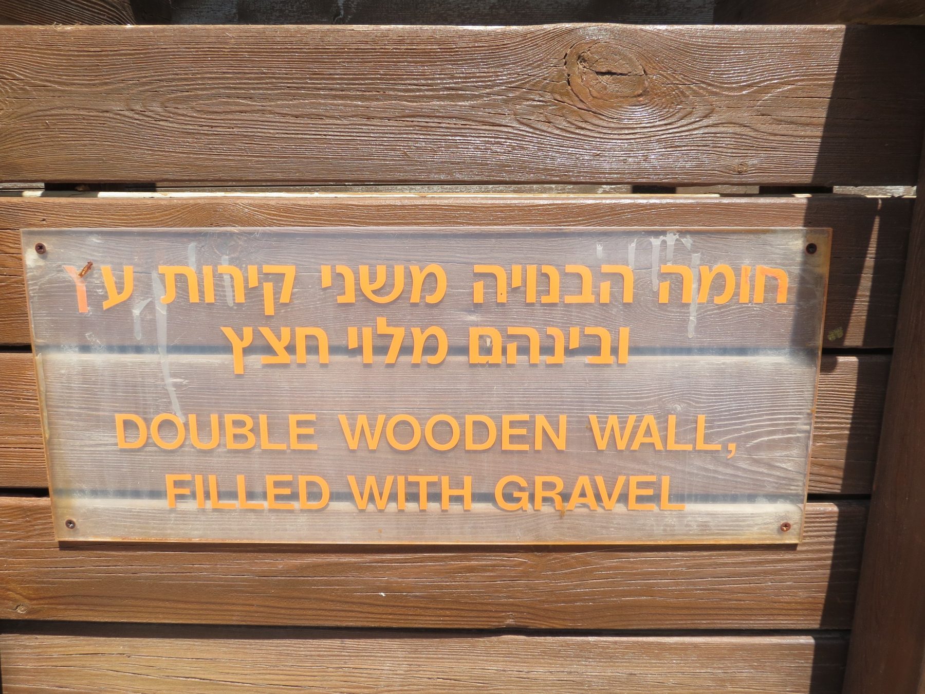 Museum model of the double wooden wall construction with interpretive bilingual label explaining the gravel-filled timber-frame design.