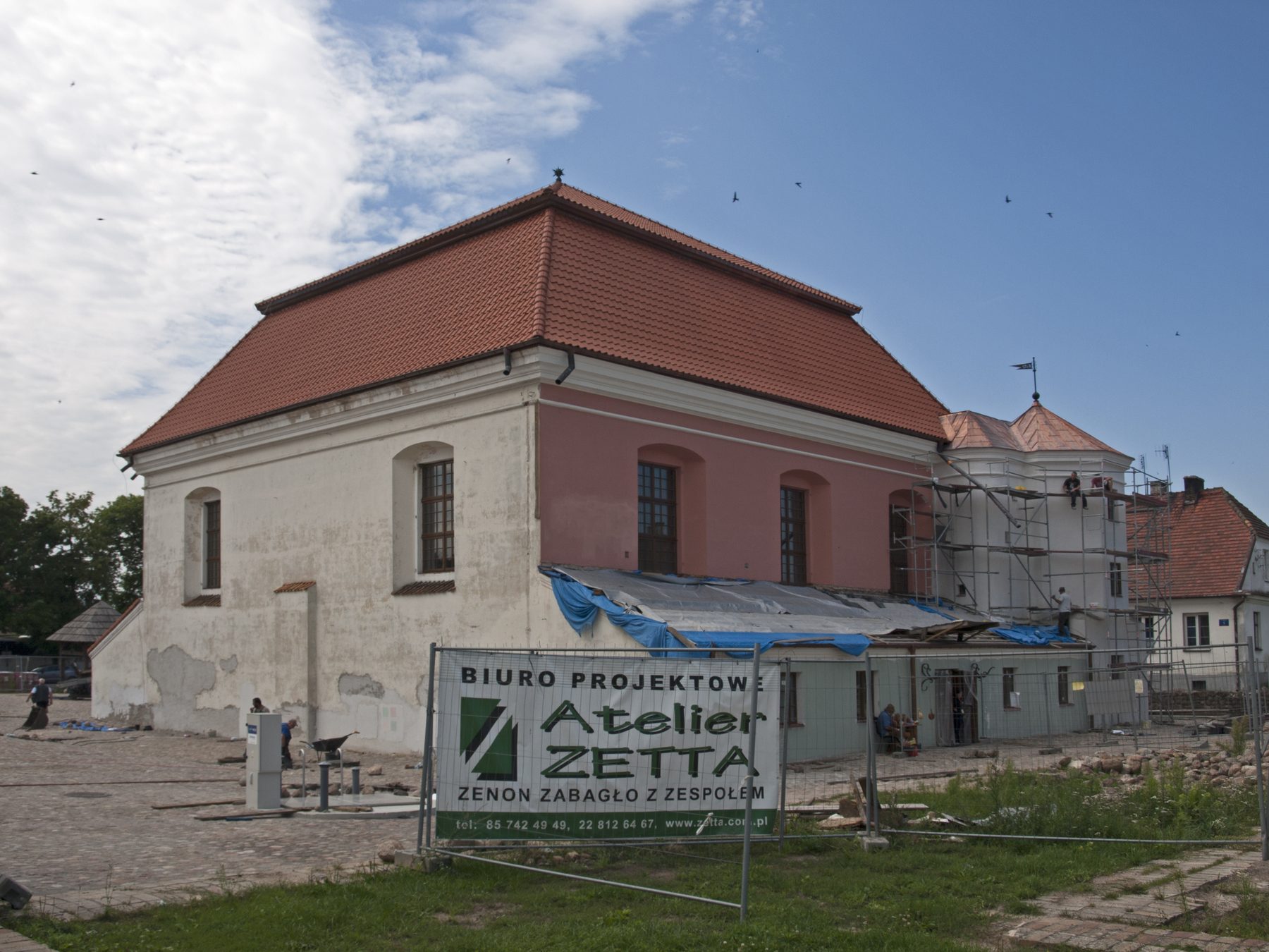 Fortress-like masonry exterior of the Tykocin Synagogue with thick stone walls, corner turrets and tiled roof, during restoration work.