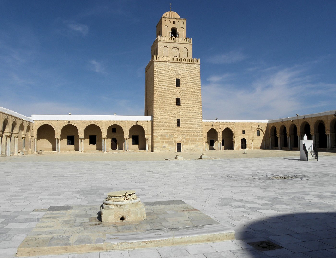 Sunlit stone courtyard with arched porticoes and a square minaret.